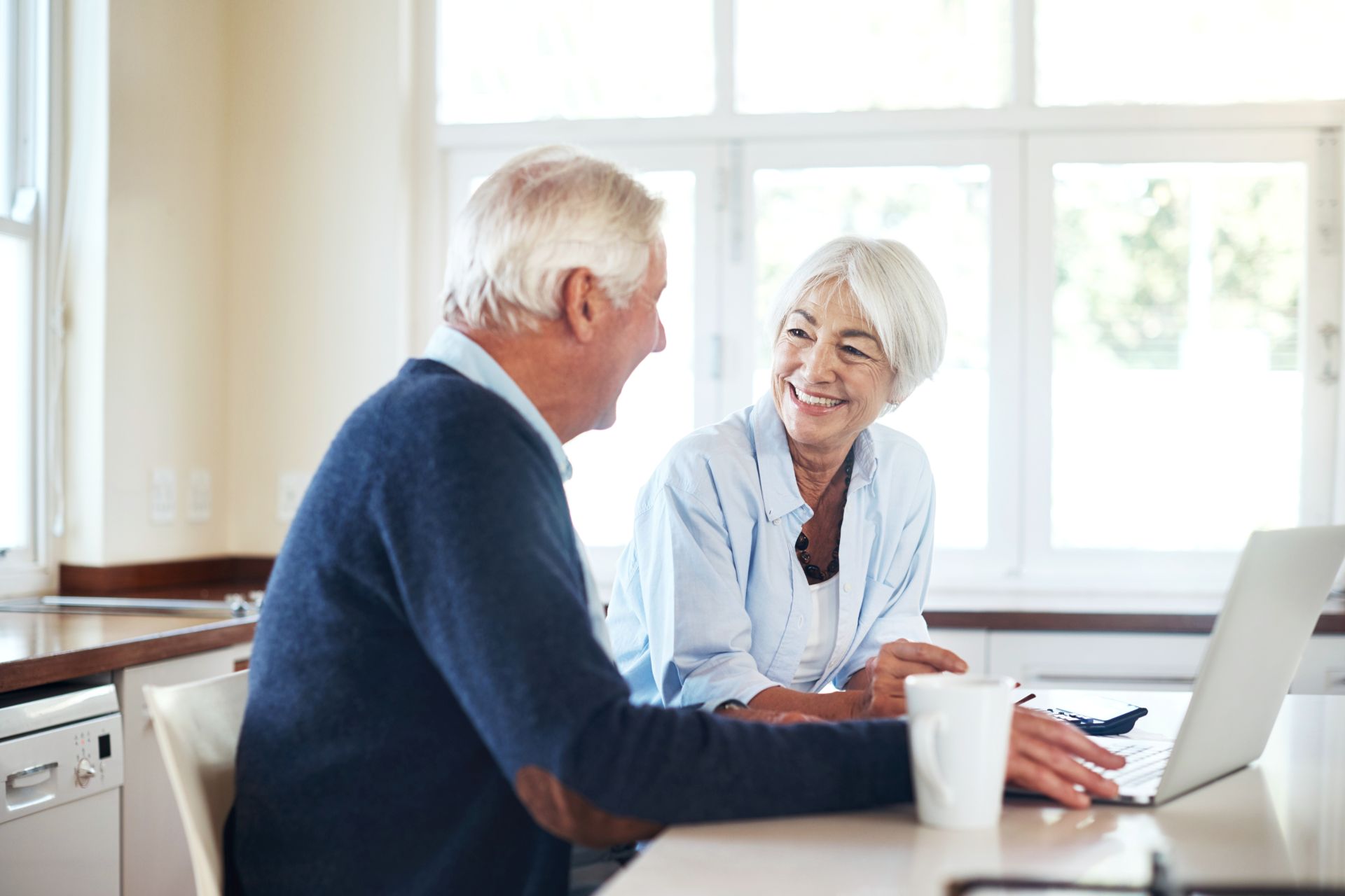 An elderly couple sitting in their kitchen smiling and looking at a laptop screen